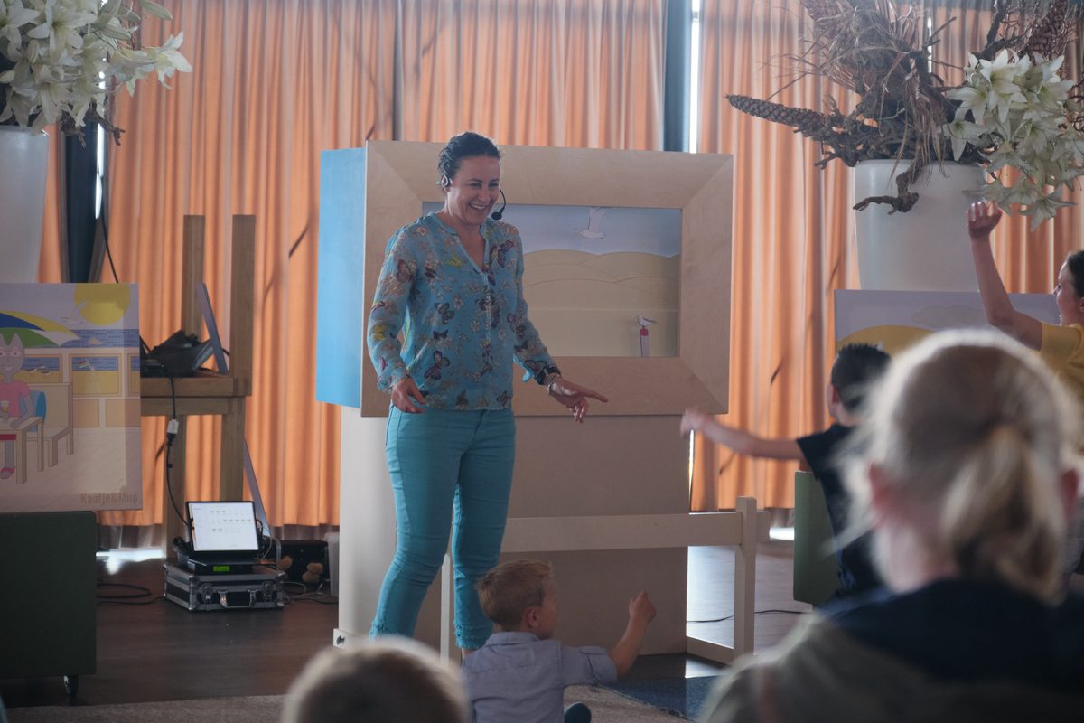 Carolien staat lachend bij de poppenkast en de kinderen kijken en steken hun hand op